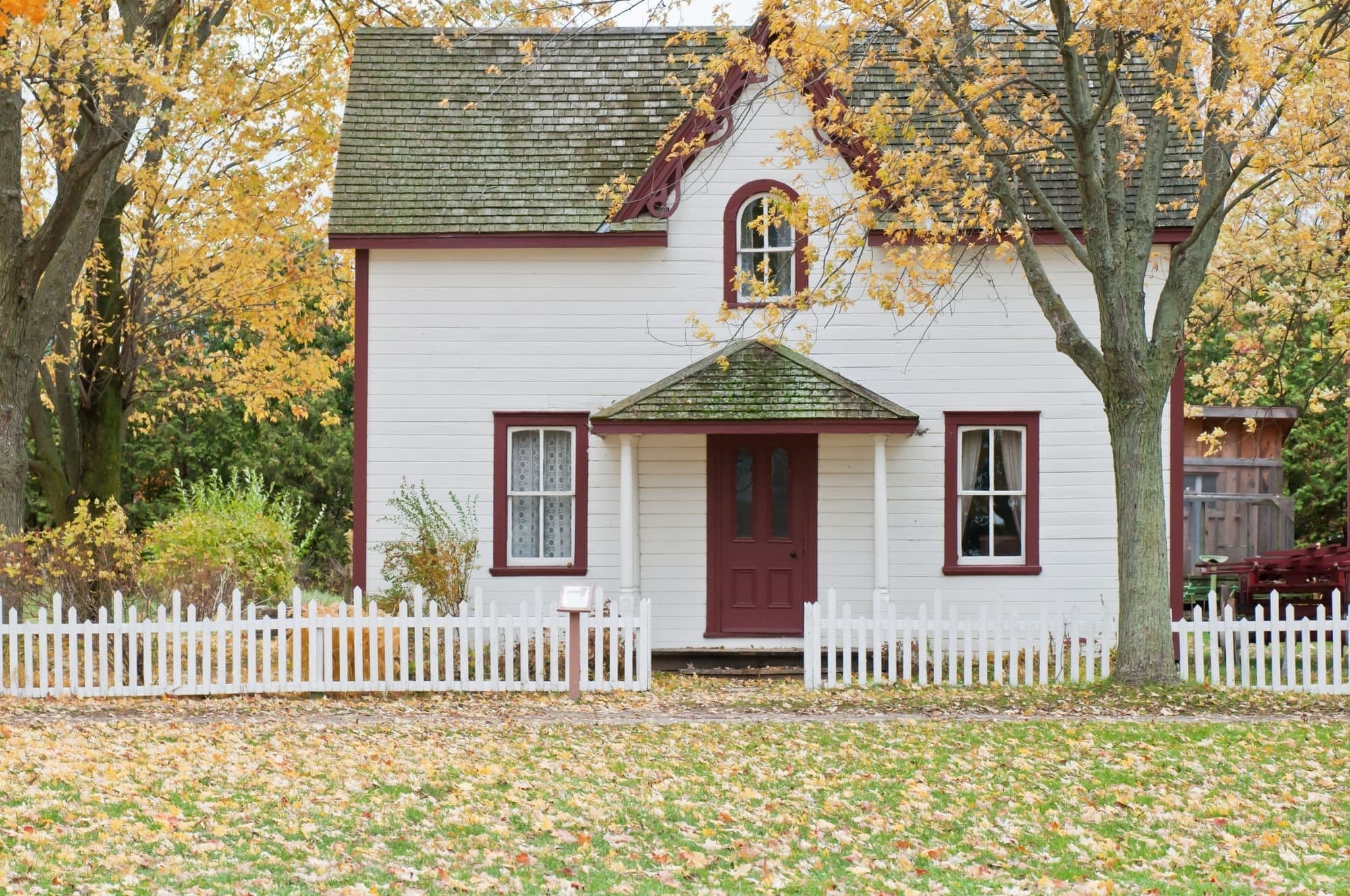 White 1800s clapboard house with a picket fence and autumn leaves